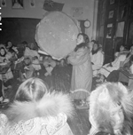 Black and white photograph of a woman playing a drum in a large building with children and adults sitting around her